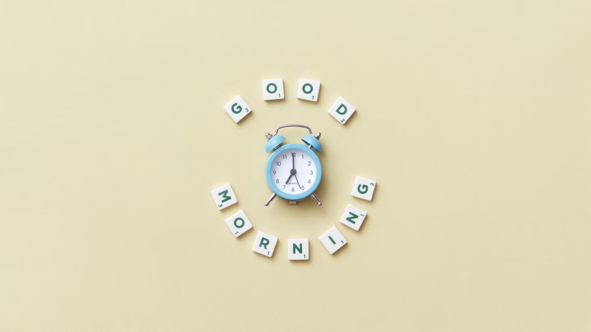 Aesthetic flat lay with a blue alarm clock surrounded by scrabble tiles spelling 'Good Morning' on a pastel background.