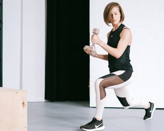 A young woman performing lunges with dumbbells in a modern indoor gym setting.